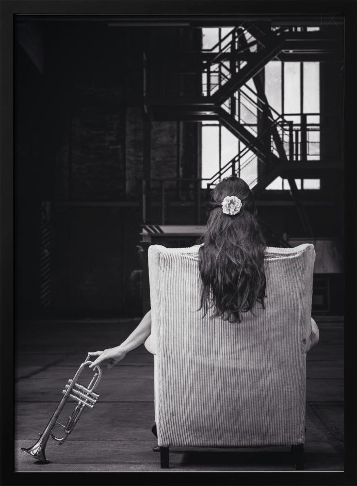 A black and white photograph of a woman seen from behind, sitting in a large, light-colored corduroy armchair. She has long, dark hair with a white flower pinned in it. Her left hand holds a trumpet, resting it on the dark floor of what appears to be an industrial warehouse with a metal staircase in the background. Wall Art