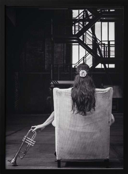 A black and white photograph of a woman seen from behind, sitting in a large, light-colored corduroy armchair. She has long, dark hair with a white flower pinned in it. Her left hand holds a trumpet, resting it on the dark floor of what appears to be an industrial warehouse with a metal staircase in the background. Wall Art