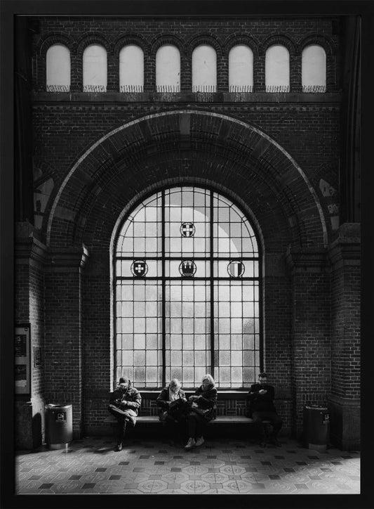 A dramatic black and white photograph of a large, brick-walled waiting area, possibly a train station. Four people are seated on a long bench beneath a massive arched window, which dominates the scene. The lighting creates a moody, contemplative atmosphere, highlighting the grandeur of the architecture. Artwork