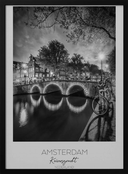 A black and white poster of Amsterdam's Keizersgracht canal at night. An arched bridge with glowing lights is the centerpiece, its reflection shimmering on the dark water. Bicycles are parked along the bridge and in the foreground on a cobblestone path. Classic canal houses and trees line the background under a dramatic, cloudy sky. The image is framed with text below reading 'AMSTERDAM Keizersgracht NEDERLAND'. Artwork