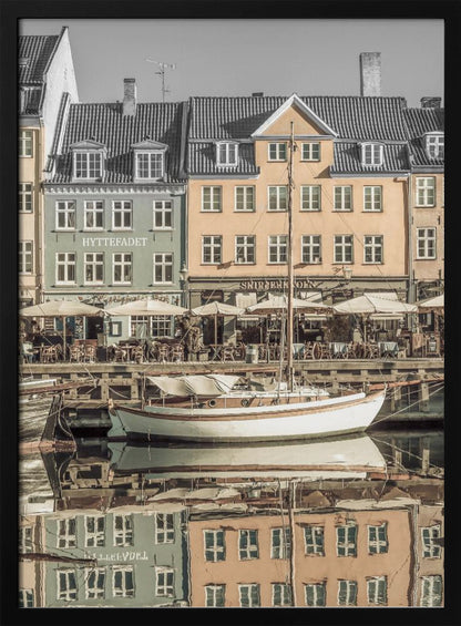 A scenic photograph of a European harbor with colorful buildings reflected in the water. A white sailboat is moored in the foreground in front of a pale green and a yellow gabled building. The image has a selective color effect, with the building facades in muted tones and the sky and roofs in black and white. Artwork