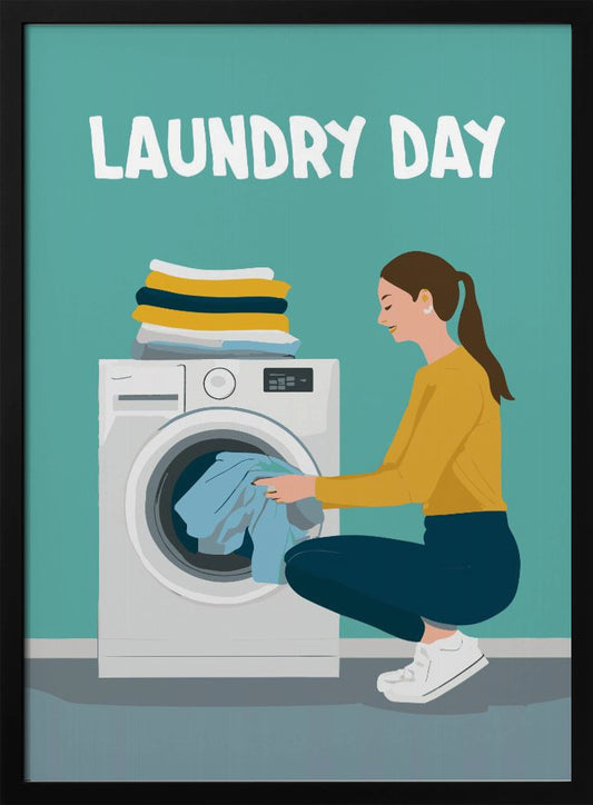 A framed illustration poster with the words 'LAUNDRY DAY' in white at the top. A woman with brown hair in a ponytail, wearing a yellow top and blue pants, crouches to load blue clothes into a white front-loading washing machine. A stack of folded laundry sits atop the machine against a teal background. Print