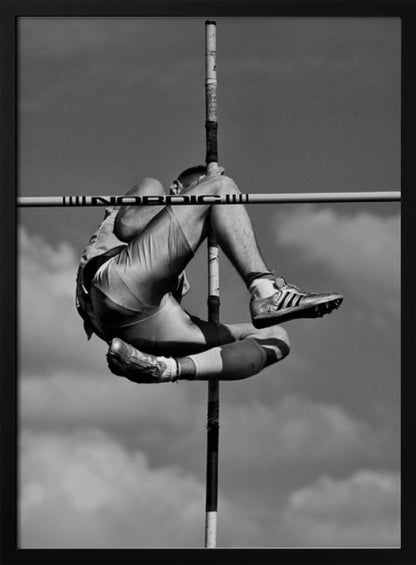 A dramatic black and white, low-angle photograph of a male athlete in mid-air during a pole vault. He is arching his back and bending his legs to clear the horizontal bar, with the vertical pole bisecting the frame. The background is a partly cloudy sky. Wall Art