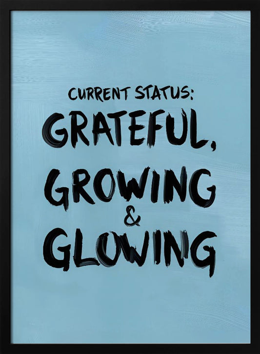 A motivational poster with a light blue background and a thin black frame. The text 'CURRENT STATUS: GRATEFUL, GROWING & GLOWING' is written in a bold, black, handwritten brush font. Decor