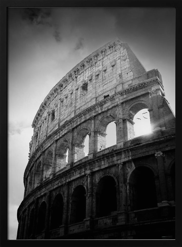 A dramatic, low-angle black and white photograph of the ancient Roman Colosseum. The sun shines brightly through one of the upper archways, creating a lens flare and silhouetting a few birds in flight against the cloudy sky. Poster