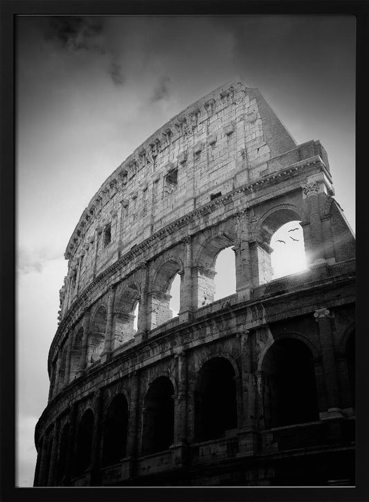 A dramatic, low-angle black and white photograph of the ancient Roman Colosseum. The sun shines brightly through one of the upper archways, creating a lens flare and silhouetting a few birds in flight against the cloudy sky. Poster