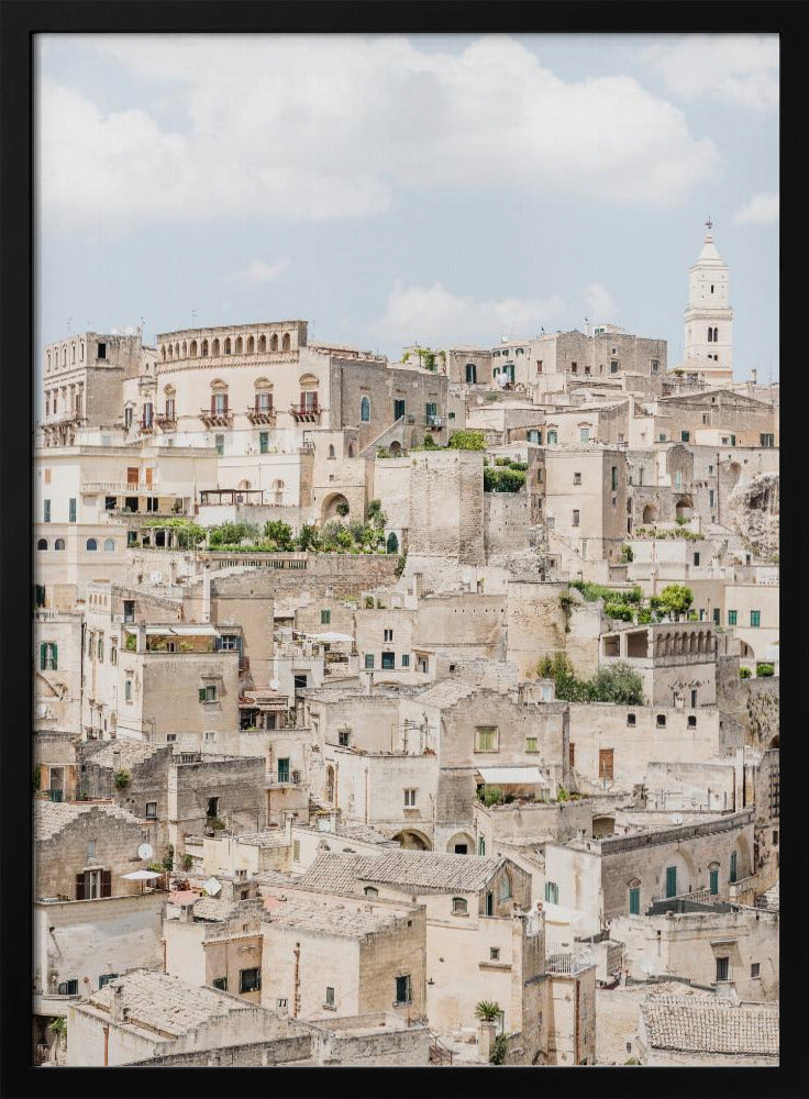 A vertical photograph of the ancient, stone city of Matera, Italy, with its dense collection of beige buildings and tiled roofs built up a hillside under a pale blue sky. A white bell tower stands out against the skyline. Poster