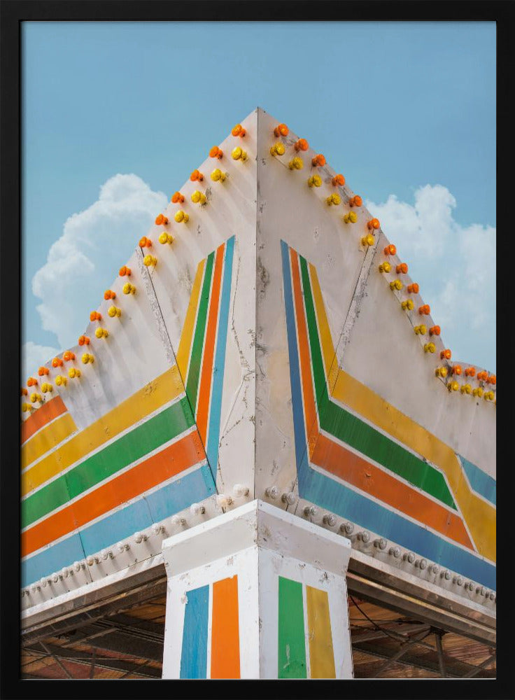 A low-angle photograph of the corner of a retro-style building against a bright blue sky with white clouds. The white facade is painted with bold, colorful stripes in orange, yellow, green, and blue, and the angled roofline is decorated with rows of unlit yellow and orange carnival lights. Print