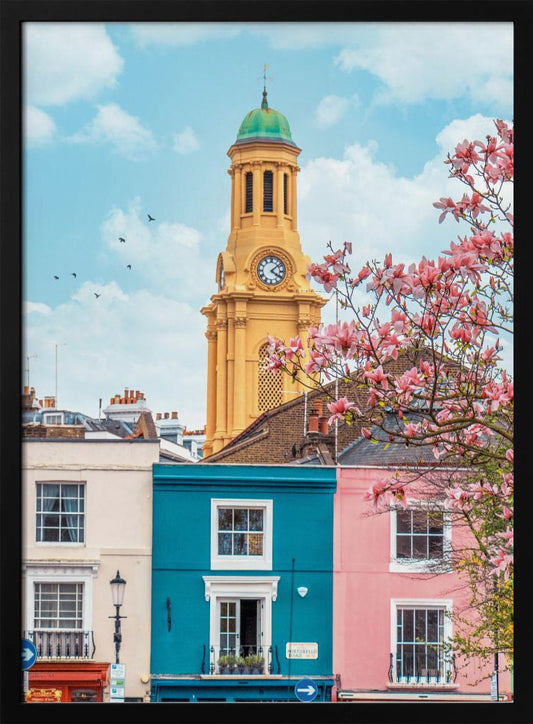 A vibrant city street scene on a sunny day, featuring a row of colorful houses in teal, pink, and off-white. A striking yellow clock tower with a green dome stands tall against a bright blue sky, with a blooming pink magnolia tree in the foreground. Print
