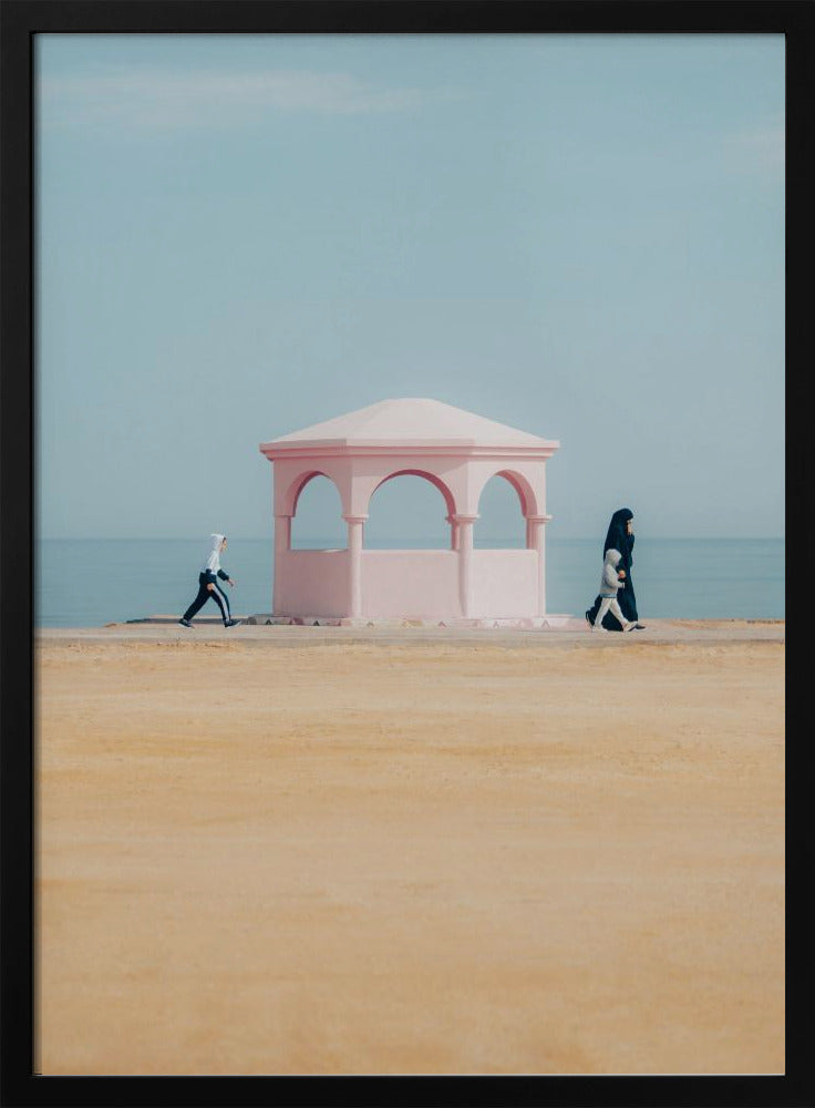 A minimalist photograph of a coastal scene with a pastel color palette. A pink arched gazebo sits on a promenade with the blue sea and sky in the background. A large expanse of beige sand is in the foreground. Two figures in hijabs walk in opposite directions on either side of the gazebo, one with a small child. Decor