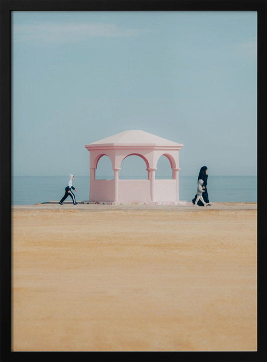 A minimalist photograph of a coastal scene with a pastel color palette. A pink arched gazebo sits on a promenade with the blue sea and sky in the background. A large expanse of beige sand is in the foreground. Two figures in hijabs walk in opposite directions on either side of the gazebo, one with a small child. Decor