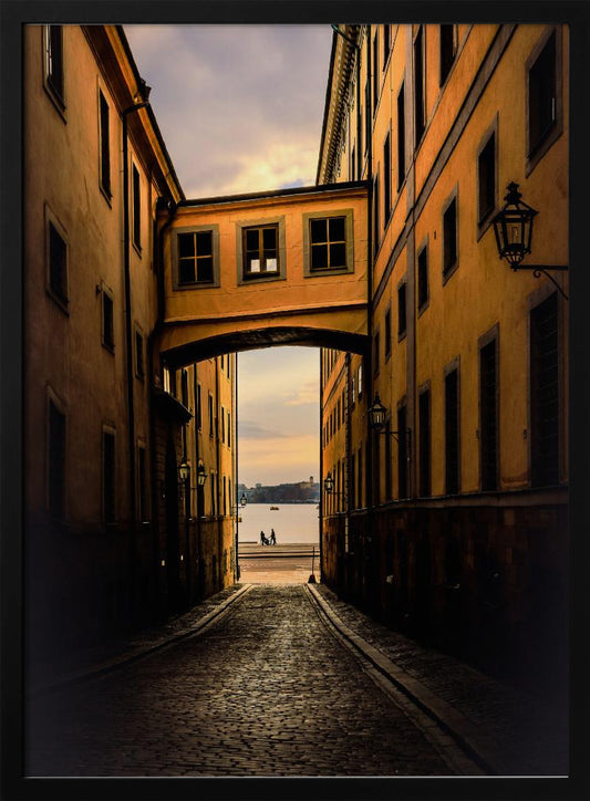 A perspective view down a narrow, shadowy cobblestone alley in an old European city, framed by warm, ochre-colored buildings. An arched walkway connects the buildings overhead, and the alley opens to a view of a tranquil body of water at sunset, with two small figures silhouetted by the shore. Decor