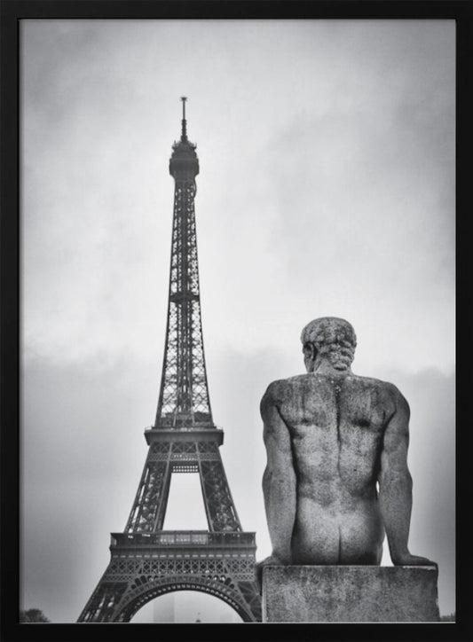 A black and white photograph capturing a contemplative scene in Paris, with the back of a seated, muscular male stone statue in the foreground looking towards the iconic Eiffel Tower which stands tall against a cloudy sky. Print