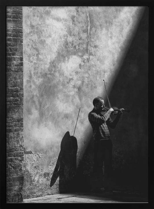 A dramatic black and white photograph of a man playing the violin, standing in a corner against a weathered wall. He is bathed in a sharp diagonal ray of light, casting a long, dark shadow behind him. Artwork