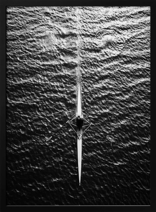 A dramatic black and white overhead photograph of a lone rower in a sleek racing shell, cutting a straight path through dark, textured water. The high-contrast lighting highlights the ripples on the water's surface and the rower's focused form. Artwork