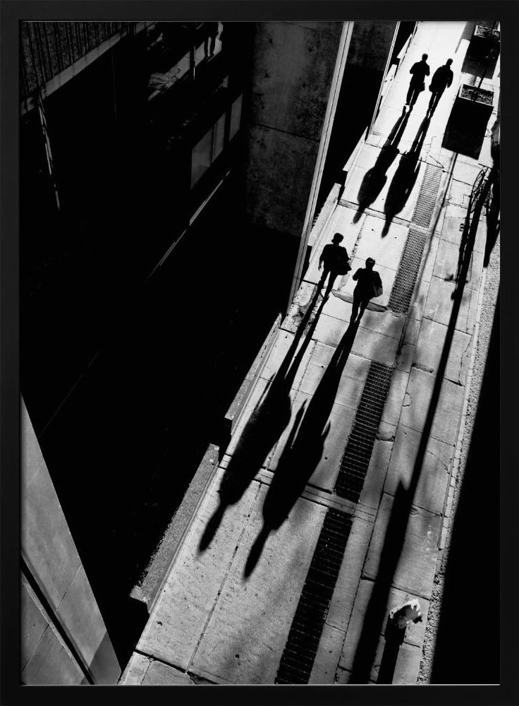 A dramatic high-angle, black and white photograph of several people walking on a sunlit sidewalk, casting long, stark shadows that stretch diagonally across the frame, contrasting with the deep black shadows of the adjacent building. Decor