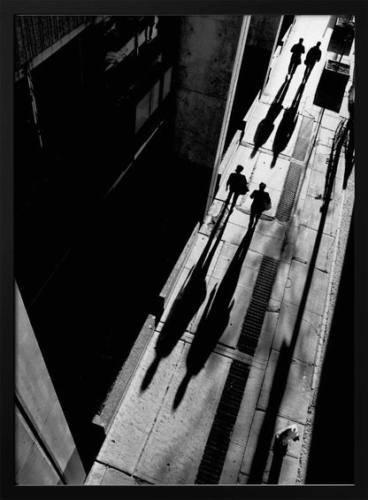 A dramatic high-angle, black and white photograph of several people walking on a sunlit sidewalk, casting long, stark shadows that stretch diagonally across the frame, contrasting with the deep black shadows of the adjacent building. Decor