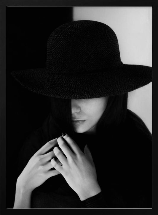 A mysterious black and white portrait of a woman wearing a large, black, wide-brimmed hat that casts a deep shadow over her eyes. Her hands are clasped at her chest, and the background is split vertically into black and white sections. Artwork