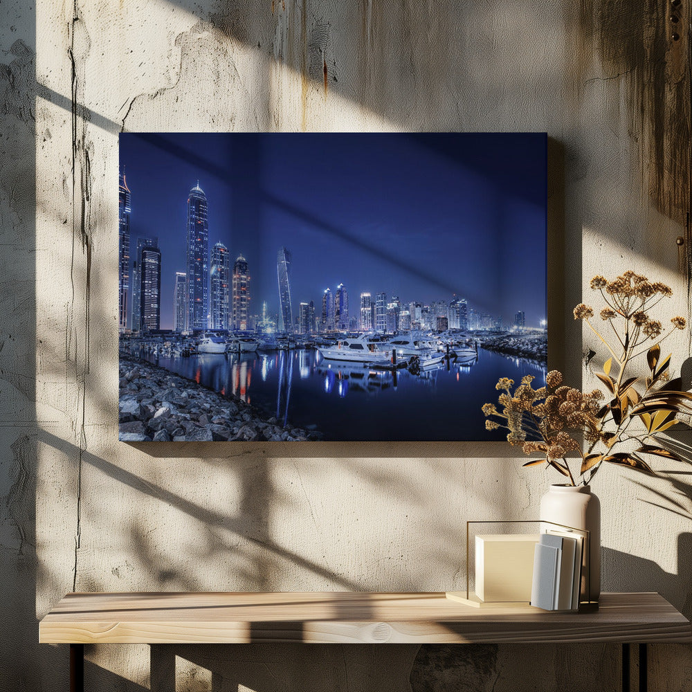 A stunning nighttime photograph of the Dubai Marina skyline, with illuminated skyscrapers towering against a deep blue sky. In the foreground, luxury yachts are moored in the calm harbor, their white hulls and the city's lights creating vibrant reflections on the water's surface. The shot is taken from a rocky embankment. Decor