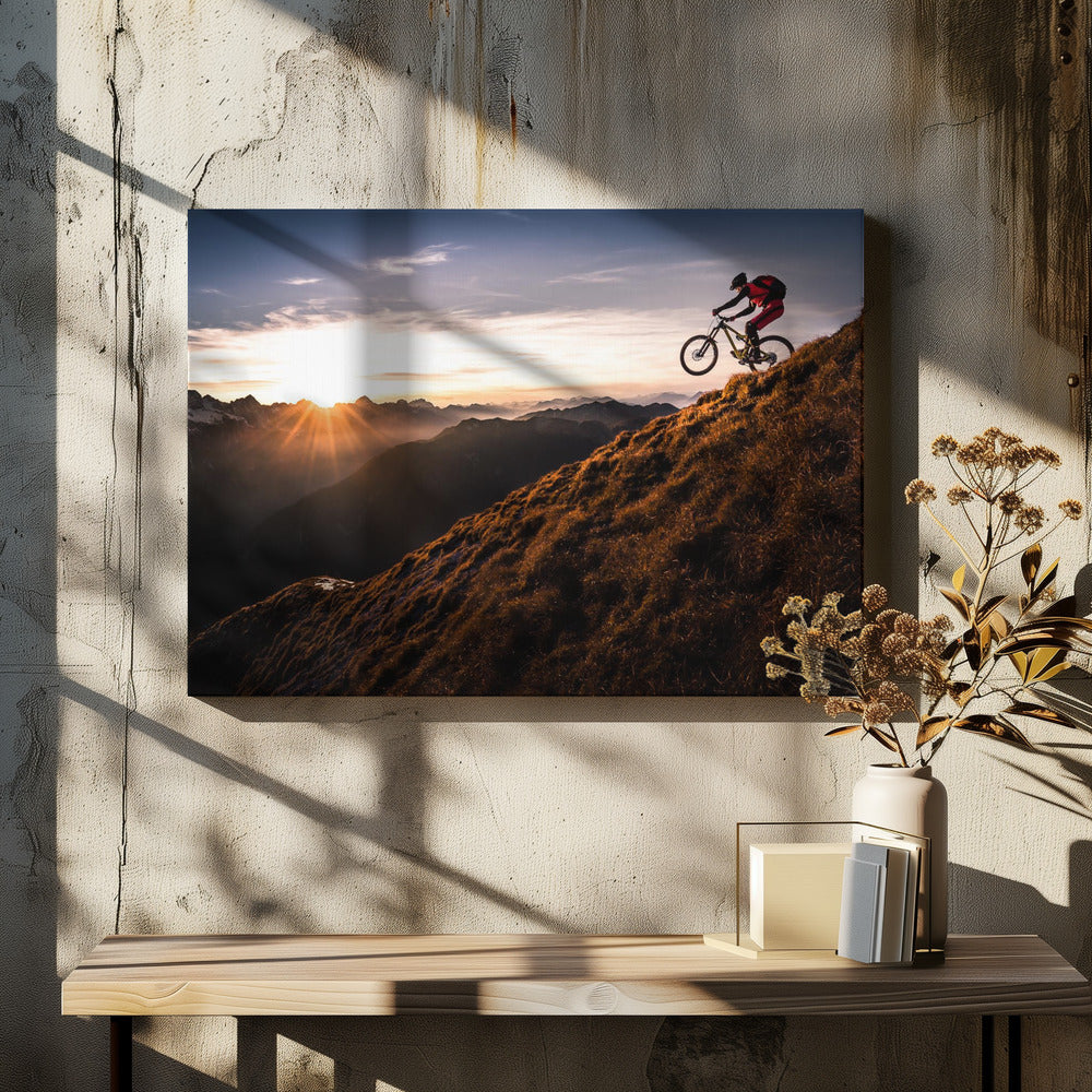 A mountain biker in mid-air, jumping down a steep grassy mountain ridge at sunrise. The sun is low on the horizon, casting a golden glow and long sunbeams over a vast, layered mountain range in the background. Poster