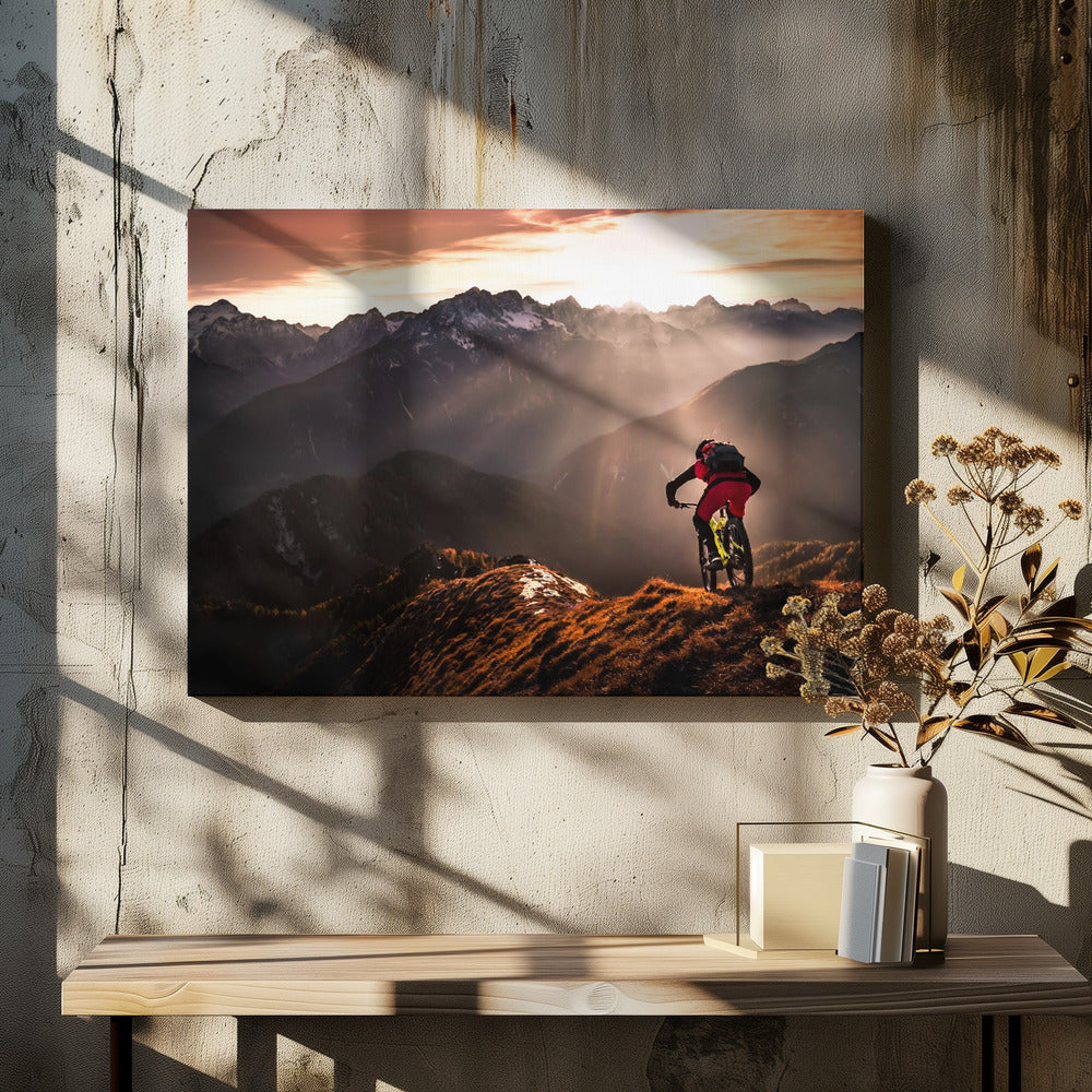 A mountain biker in red and black gear pauses on a grassy, sunlit ridgeline, looking out at a vast mountain range with snow-capped peaks as the sun sets behind them, casting dramatic rays of light through the valleys. Poster