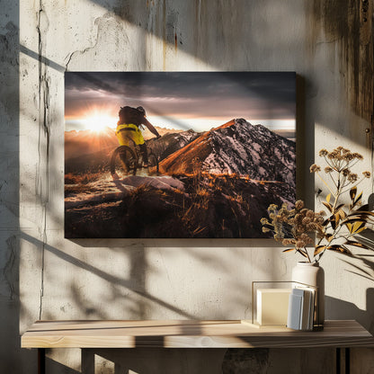 A mountain biker seen from behind rides along a narrow, snowy trail on a mountain ridge during a dramatic sunset, with the sun flaring brightly and a vast, snow-capped mountain range in the background. Decor