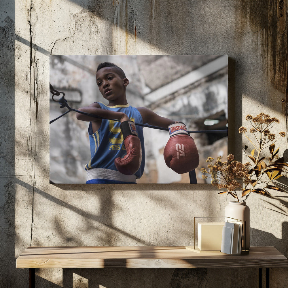 A young Black boy in a blue jersey and red boxing gloves leans pensively against the ropes of a boxing ring, looking away with a thoughtful expression against a blurred, weathered background. Wall Art
