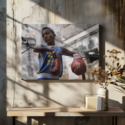 A young Black boy in a blue jersey and red boxing gloves leans pensively against the ropes of a boxing ring, looking away with a thoughtful expression against a blurred, weathered background. Wall Art