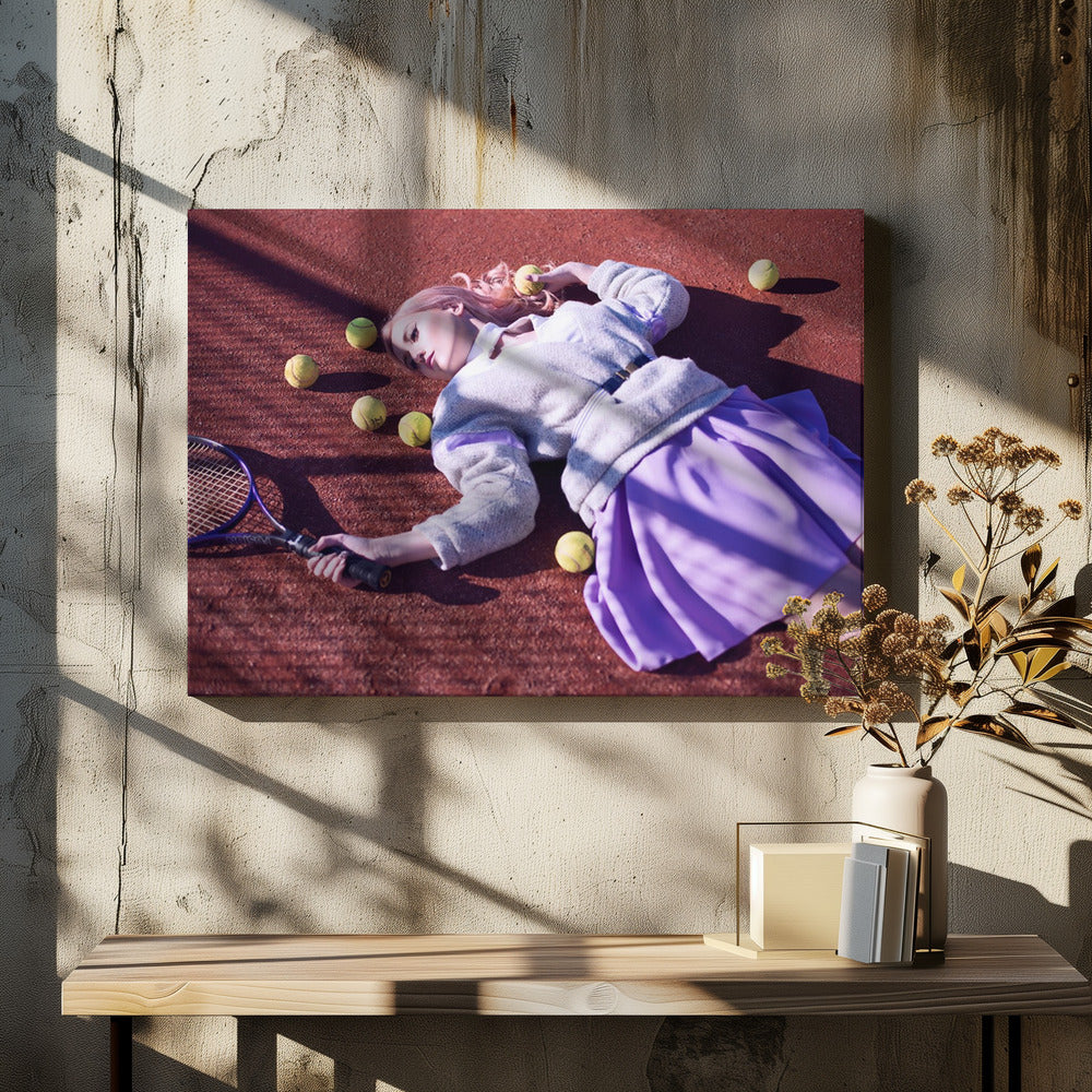 A high-angle shot of a young woman with pink hair lying on a red clay tennis court, surrounded by yellow tennis balls. She wears a grey sweater, a white collared shirt, and a purple pleated skirt, and holds a tennis racket in one hand. The scene is framed with a silver border. Wall Art
