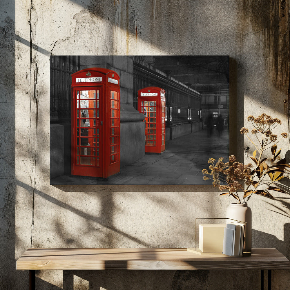 A selective color photograph of a city street at night, framed in silver. Two iconic red telephone booths are the only objects in color, standing out against the black and white background of the cobblestone sidewalk and iron fence. Artwork
