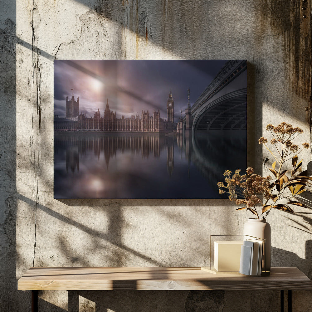 A dramatic, atmospheric photograph of the Houses of Parliament and Big Ben in London, reflected in the still waters of the River Thames. A bright sun breaks through dark, moody clouds, casting a warm light on the scene, with a stone bridge visible to the right. The image is enclosed in a silver frame. Print
