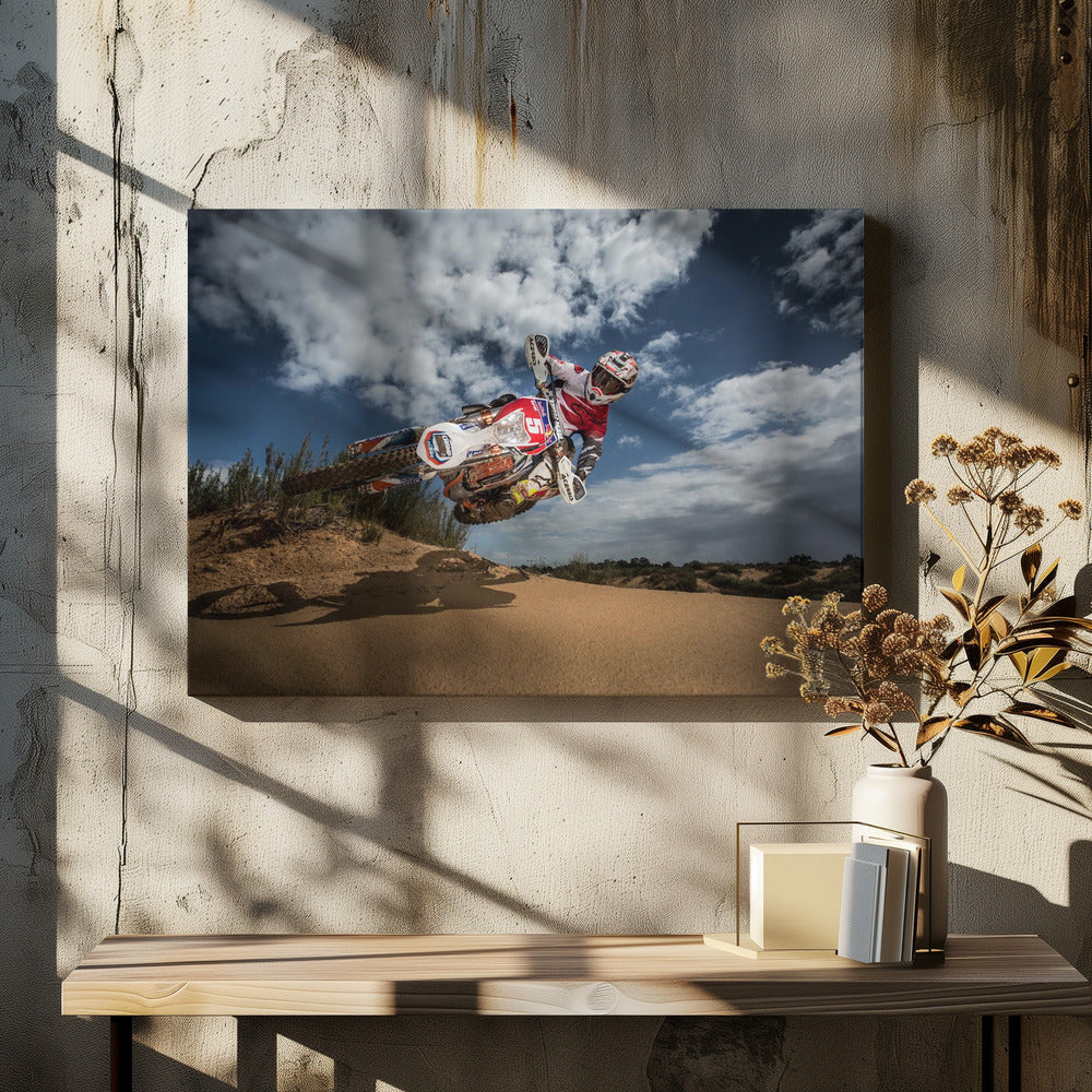 A low-angle action shot of a motocross rider on a dirt bike catching major air over a sand dune against a cloudy blue sky. The image is presented within a silver frame. Decor