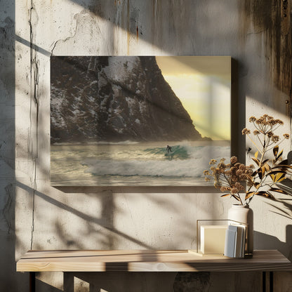 A framed photograph captures a lone surfer in a black wetsuit riding a turquoise wave. In the background, a massive, steep, snow-dusted mountain looms over the cold sea under a pale yellow, overcast sky. Print