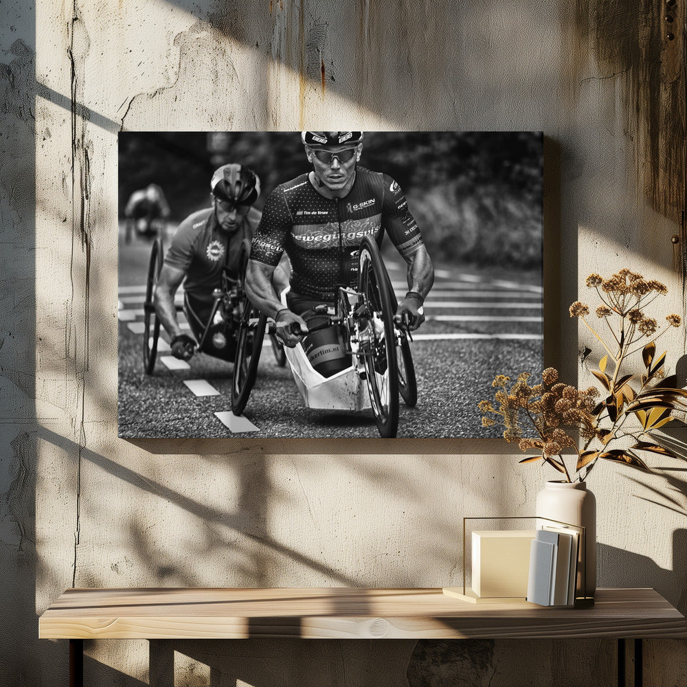 A powerful black and white photograph of two male athletes competing in a wheelchair race. The focused athlete in the foreground shows intense determination on his face, his muscular arms propelling the specialized racing wheelchair forward on an asphalt road. A second racer is visible in the background. Decor