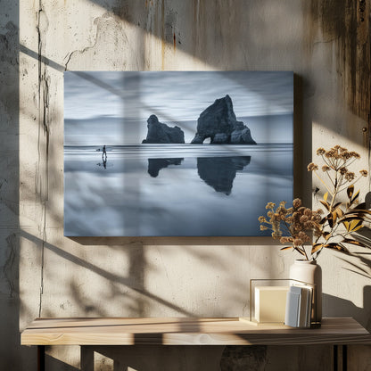 A moody, blue-toned monochromatic photograph of a lone surfer walking on a wet, reflective beach. Two large rock formations stand in the calm ocean in the background, their reflections mirrored in the water. The sky is overcast, creating a serene and atmospheric scene, all enclosed in a silver frame. Decor