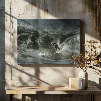 A dramatic overhead photograph of a surfer in a black wetsuit riding a white surfboard on a large, dark, foamy wave. The scene is captured in a moody, almost monochromatic tone and is presented within a silver picture frame. Artwork