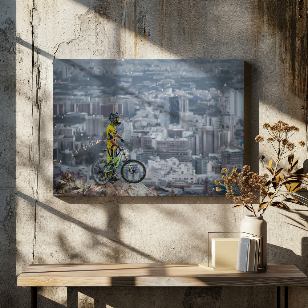 A mountain biker in a yellow and blue uniform stands with a green and white full-suspension bike on a rocky precipice, looking out over a vast, hazy cityscape that stretches into the distance. Print