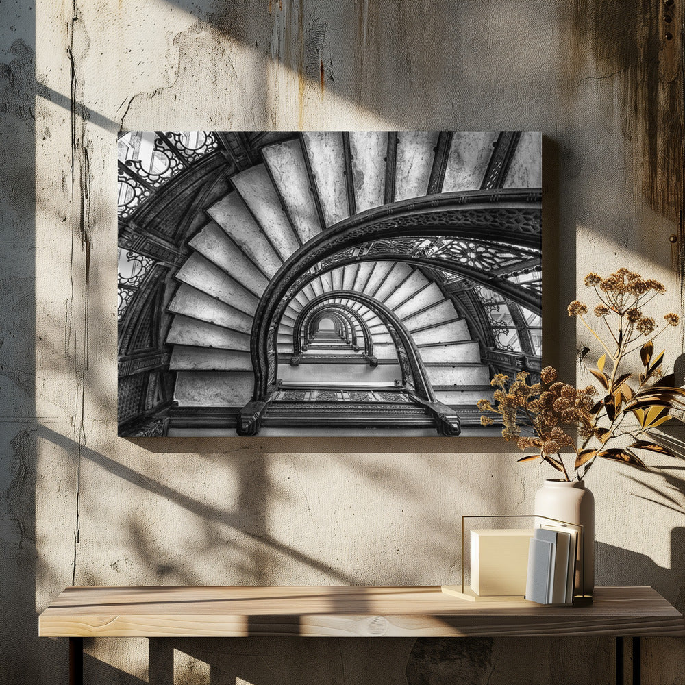 A dramatic black and white photograph looking up from the bottom of an ornate spiral staircase. The dark, intricate ironwork of the railings contrasts with the light stone steps, creating a mesmerizing, repeating pattern that spirals towards a bright light at the top. The image is presented within a silver-colored frame. Decor