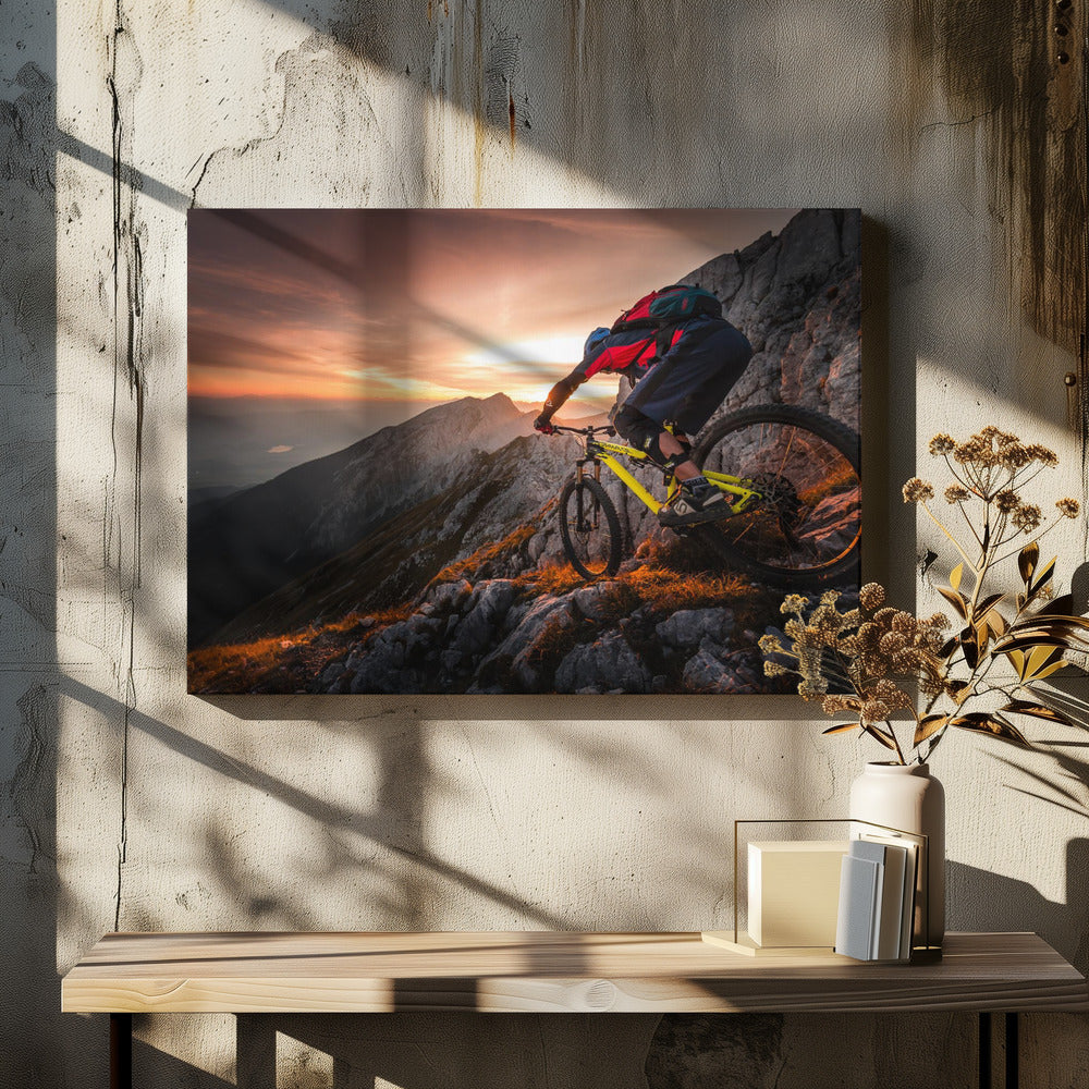 A mountain biker in full gear rides a yellow bike down a narrow, rocky mountain ridge at sunset. The sky is filled with the warm colors of the setting sun, which illuminates the distant mountain peaks. Artwork