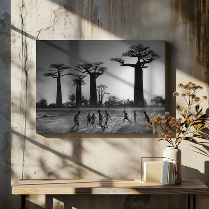 A framed, black and white photograph capturing a vibrant scene of several children playing soccer on a dirt field. In the background, the iconic silhouettes of large Baobab trees stand tall against a bright, hazy sun, casting long shadows of the players across the ground. Artwork