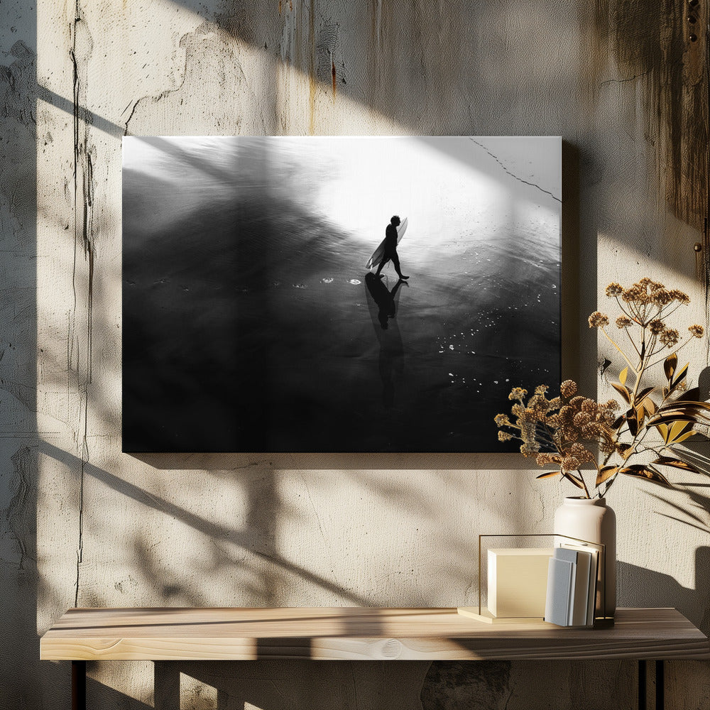 A high-contrast, black and white photograph of a surfer silhouetted against the bright reflection of the sun on the water. The surfer is carrying a surfboard and walking along the wet sand, leaving footprints behind. Their long shadow is cast in front of them on the dark, glistening sand. Print
