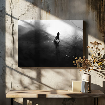 A high-contrast, black and white photograph of a surfer silhouetted against the bright reflection of the sun on the water. The surfer is carrying a surfboard and walking along the wet sand, leaving footprints behind. Their long shadow is cast in front of them on the dark, glistening sand. Print