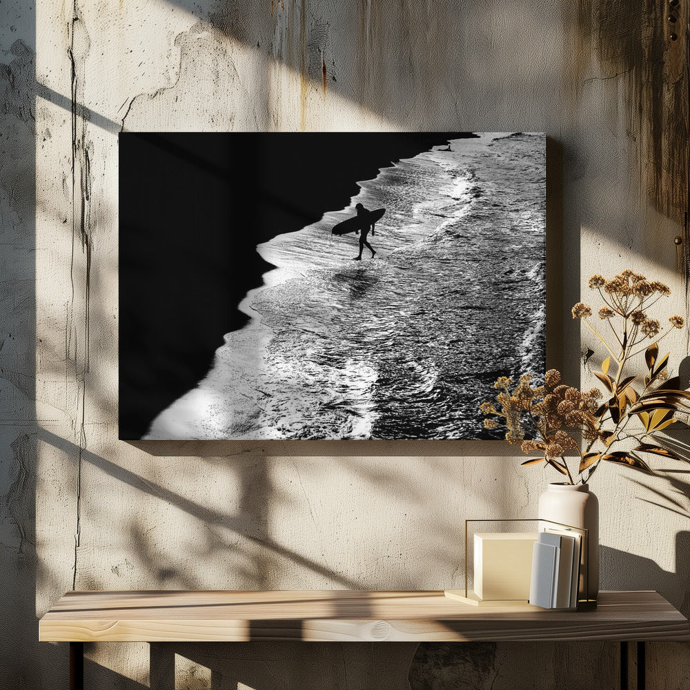 A high-contrast, black and white aerial photograph of a silhouetted surfer carrying a surfboard and walking along the shoreline. The image is divided diagonally, with the dark beach on the left and the white, foamy ocean waves on the right, creating a dramatic and graphic composition. The image is enclosed in a silver frame. Wall Art
