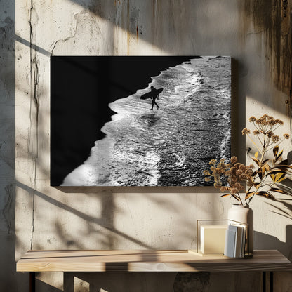 A high-contrast, black and white aerial photograph of a silhouetted surfer carrying a surfboard and walking along the shoreline. The image is divided diagonally, with the dark beach on the left and the white, foamy ocean waves on the right, creating a dramatic and graphic composition. The image is enclosed in a silver frame. Wall Art