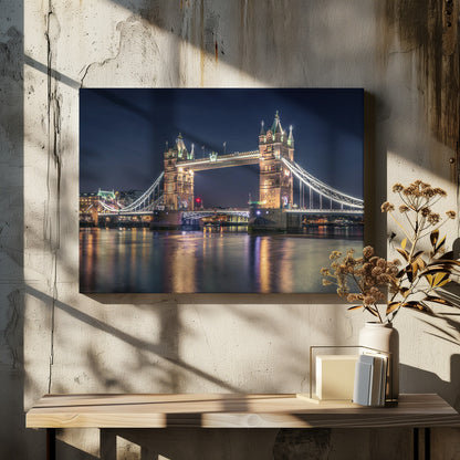 A stunning night photograph of London's iconic Tower Bridge, fully illuminated with golden and white lights that cast a shimmering reflection on the dark waters of the River Thames below, all set against a deep night sky and enclosed in a silver frame. Poster