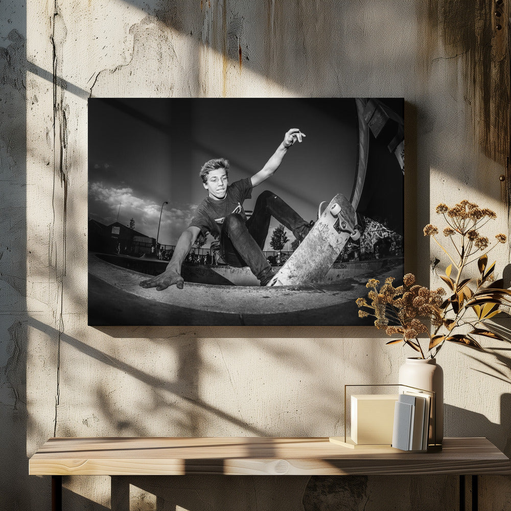 A dynamic, low-angle black and white action shot of a young skateboarder performing a trick in a concrete skatepark. He balances with one hand on the edge of the bowl, his body low to the ground as he carves along the curve, with his other arm raised in the air. Wall Art