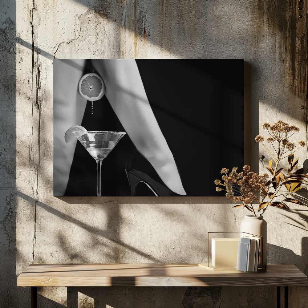 A framed, black and white photograph showing a woman's legs and a martini glass against a black background. A sliced citrus fruit is held between her thighs, with juice dripping towards the sugar-rimmed cocktail glass below. One foot is visible, wearing a black stiletto high heel. Artwork