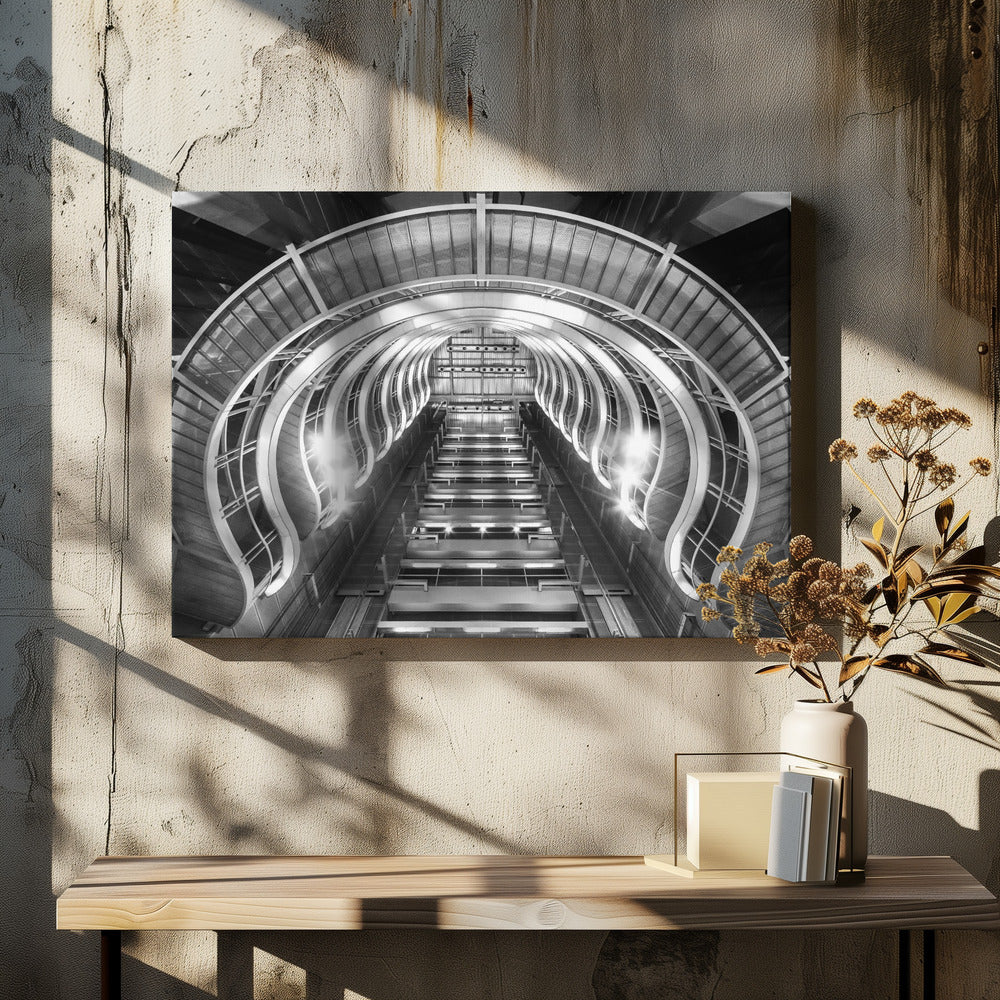 A symmetrical, low-angle black and white photograph of a futuristic escalator or grand staircase. The structure features illuminated, repeating curved arches on both sides, creating a tunnel-like perspective leading upwards. The image is presented within a silver-colored frame. Artwork