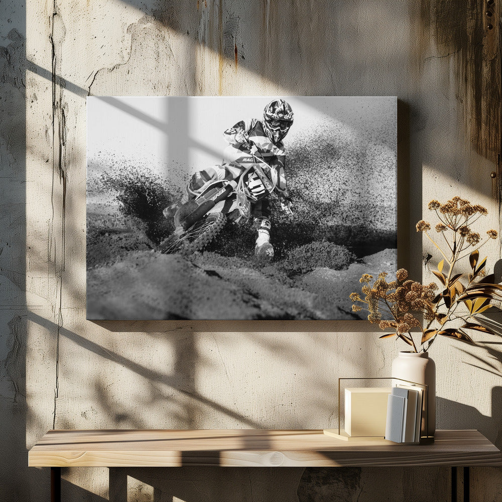 A dynamic black and white action photo of a motocross rider in full gear leaning hard into a turn, kicking up a large spray of dirt and sand from the track. The image is presented within a silver frame. Decor
