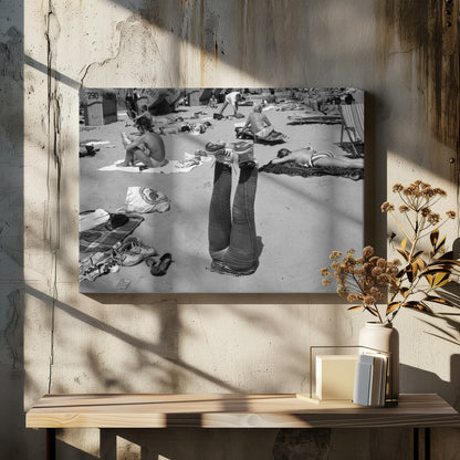 A quirky, vintage black and white photograph of a crowded beach. The central focus is a person lying on their back with their legs straight up in the air, oddly wearing jeans and sneakers. Surrounding them are numerous other beachgoers sunbathing on towels and in wicker beach chairs, creating a busy and candid summer scene. Wall Art